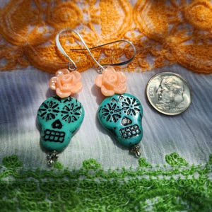 May include: Turquoise skull earrings with black detailing and peach rose accents. The earrings are shown with a coin for scale. The Day of the Dead-themed earrings feature intricate designs, showcasing the craftsmanship of the jewellery.