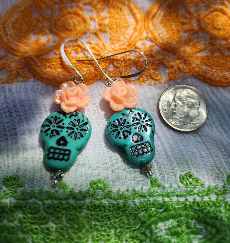 May include: Turquoise skull earrings with black detailing and peach rose accents. The earrings are shown with a coin for scale. The Day of the Dead-themed earrings feature intricate designs, showcasing the craftsmanship of the jewellery.