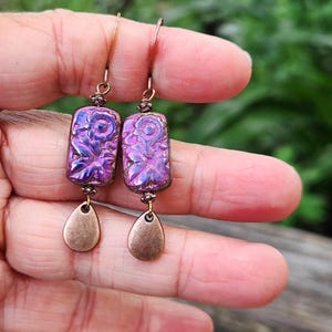 May include: A pair of dangle earrings featuring rectangular purple iridescent beads with a floral relief pattern, suspended from copper-toned ear wires. Each earring includes a small, smooth, copper-toned teardrop-shaped charm at the base.