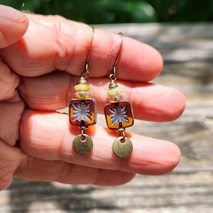May include: Dangle earrings featuring square amber-coloured glass beads with a central white starburst pattern. Each earring includes small iridescent accent beads, a brass-toned metal hook, and a small, flat, circular brass-toned metal charm at the base.