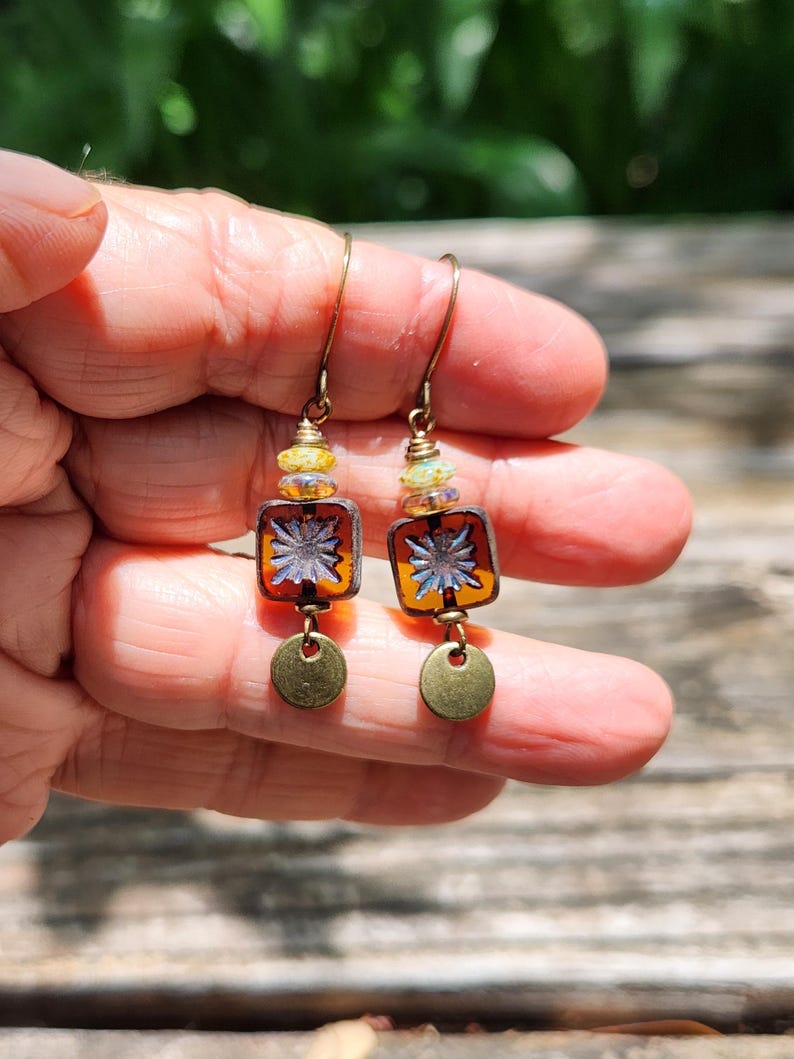 May include: Dangle earrings featuring square amber-coloured glass beads with a central white starburst pattern. Each earring includes small iridescent accent beads, a brass-toned metal hook, and a small, flat, circular brass-toned metal charm at the base.