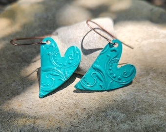May include: A pair of turquoise heart-shaped earrings with copper-colored ear wires. The earrings feature an embossed floral design. The earrings are displayed on a stone surface.