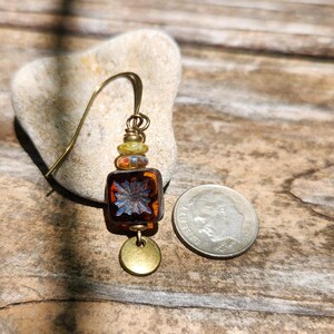 May include: A single earring featuring a dark amber square glass bead with a blue floral pattern, topped with two smaller multicoloured beads and a brass hook. A small round brass disc hangs at the bottom, next to a US dime for scale.