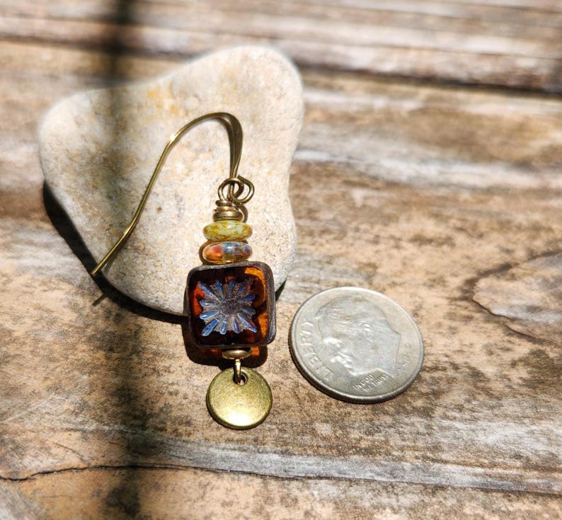 May include: A single earring featuring a dark amber square glass bead with a blue floral pattern, topped with two smaller multicoloured beads and a brass hook. A small round brass disc hangs at the bottom, next to a US dime for scale.