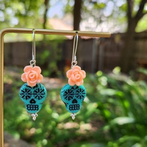 May include: Dangle earrings with turquoise skull beads, black floral patterns, and peach rose accents. The earrings hang from silver hooks. The design is inspired by the Day of the Dead, a Mexican holiday.