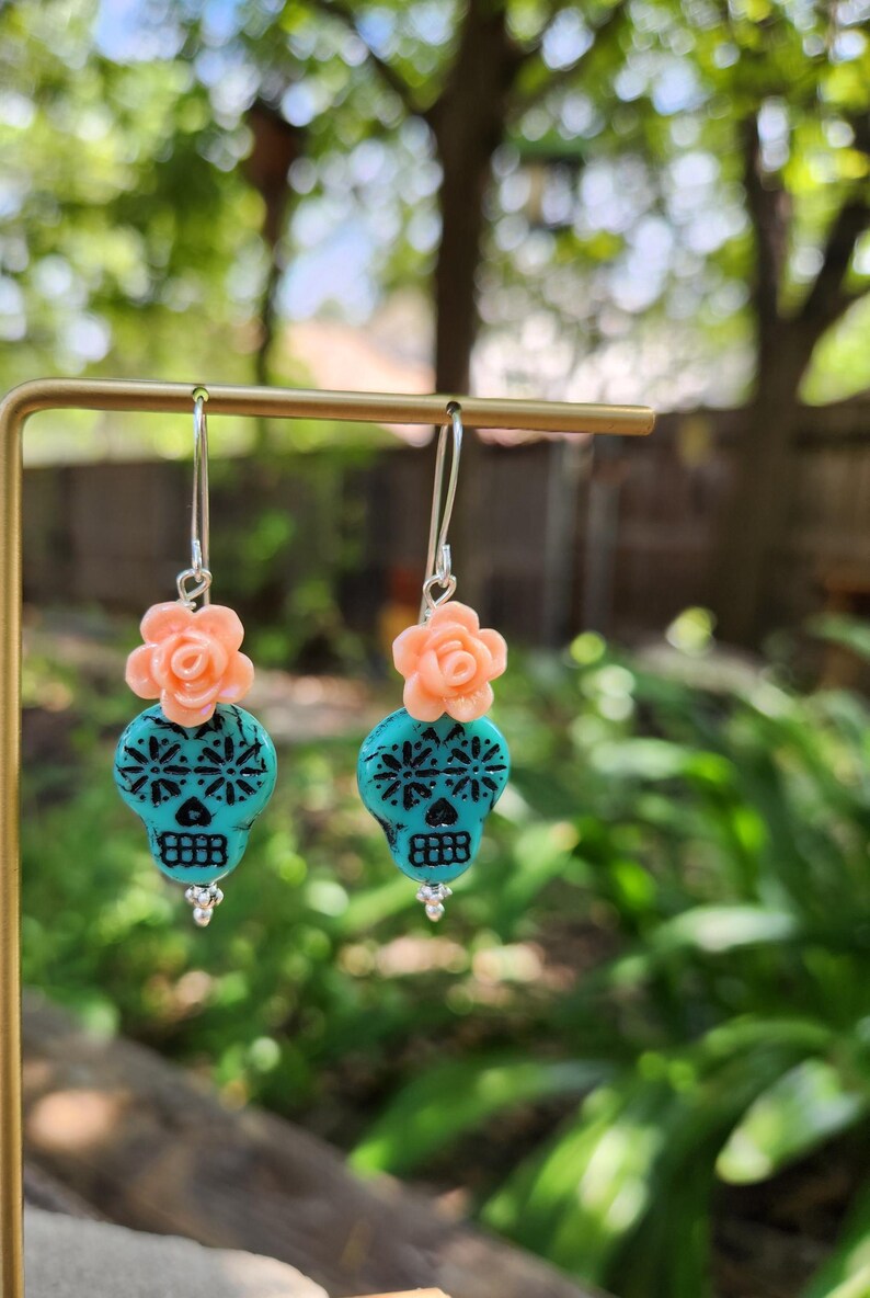 May include: Dangle earrings with turquoise skull beads, black floral patterns, and peach rose accents. The earrings hang from silver hooks. The design is inspired by the Day of the Dead, a Mexican holiday.
