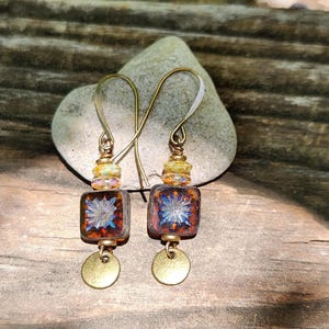 May include: A pair of drop earrings featuring square, amber-coloured glass beads with a blue starburst pattern. Each earring includes small, multi-coloured accent beads, a brass-toned metal hook, and a small, round, flat brass-toned metal charm.