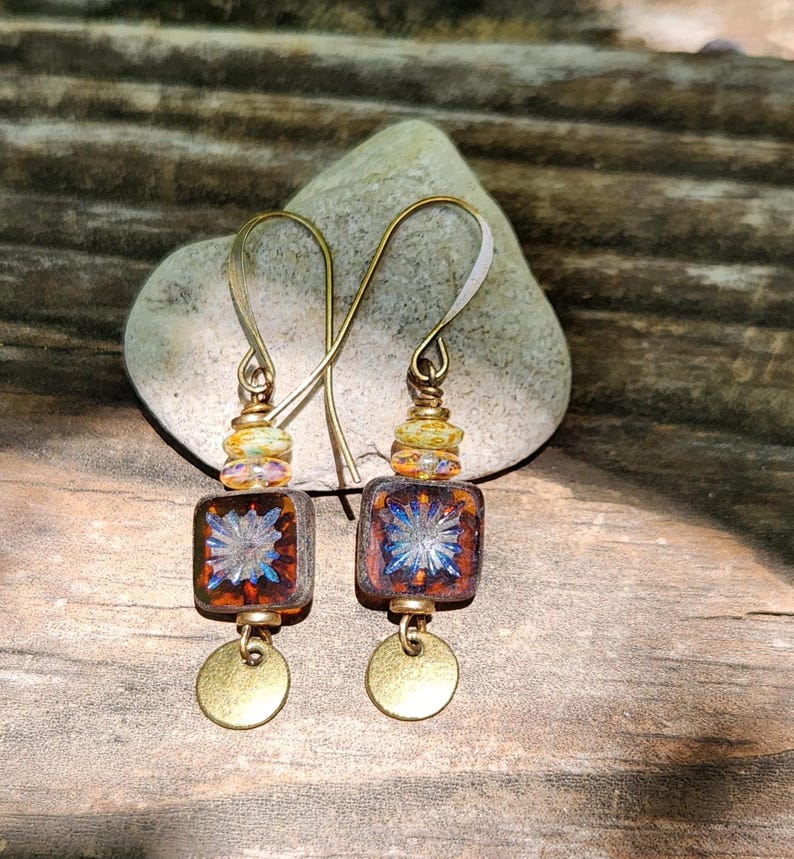 May include: A pair of drop earrings featuring square, amber-coloured glass beads with a blue starburst pattern. Each earring includes small, multi-coloured accent beads, a brass-toned metal hook, and a small, round, flat brass-toned metal charm.