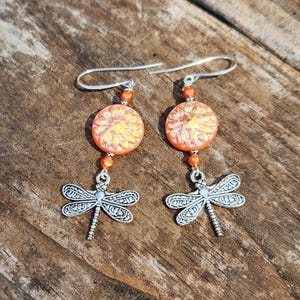 May include: A pair of dangle earrings featuring orange and yellow circular beads with a floral design. Below the beads are silver dragonfly charms. The earrings hang from silver ear wires. The background is a wooden surface.