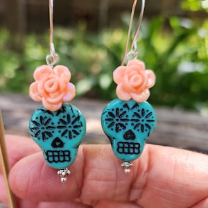May include: Dangle earrings with turquoise skull beads, black floral patterns, and peach flower accents. The earrings have silver hooks and small silver bead details. The earrings are a unique accessory.