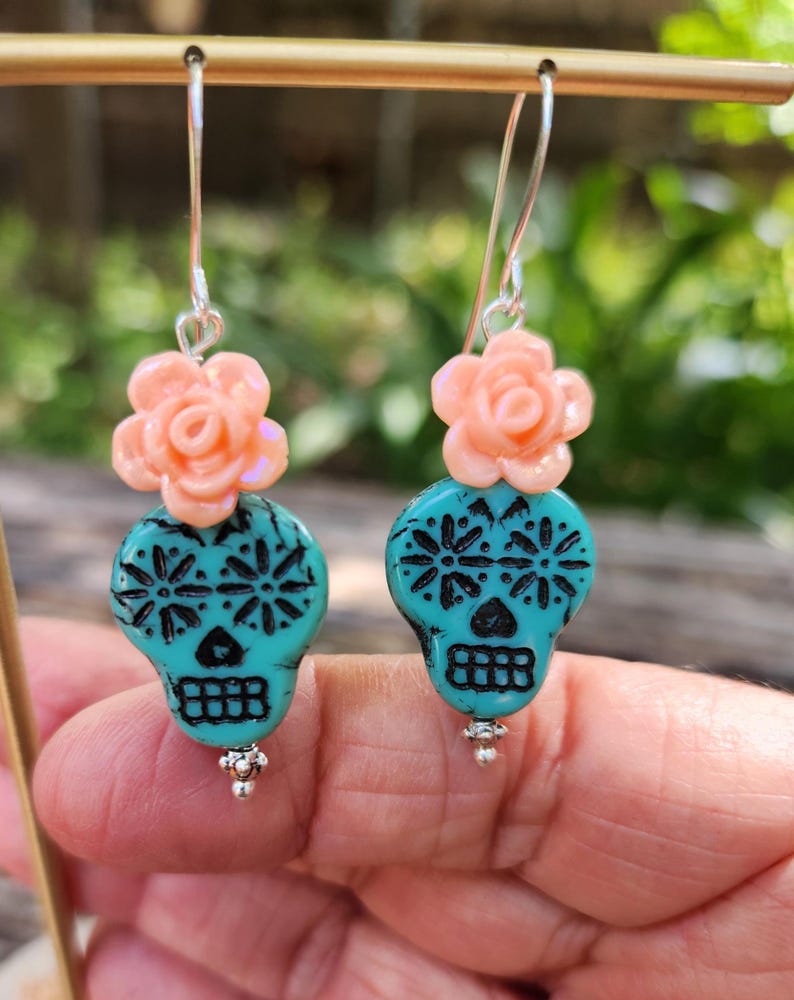 May include: Dangle earrings with turquoise skull beads, black floral patterns, and peach flower accents. The earrings have silver hooks and small silver bead details. The earrings are a unique accessory.