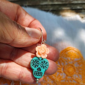 May include: A turquoise skull-shaped earring with black floral designs and a peach-coloured rose accent. The earring is suspended from a silver hook. The design is inspired by the Day of the Dead.
