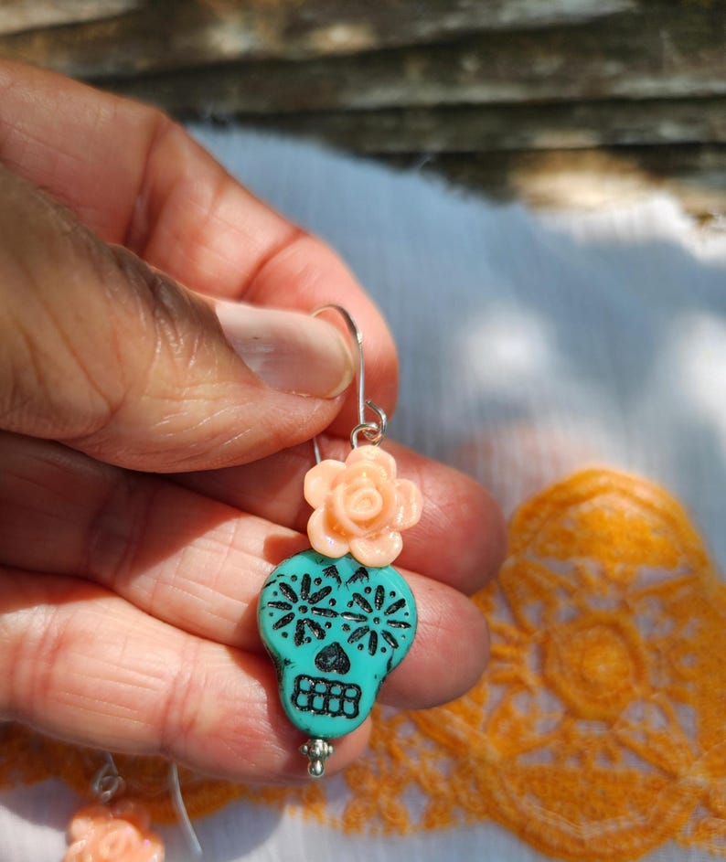 May include: A turquoise skull-shaped earring with black floral designs and a peach-coloured rose accent. The earring is suspended from a silver hook. The design is inspired by the Day of the Dead.