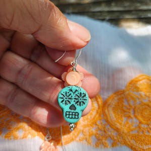 May include: Dangle earrings with turquoise skull beads and black floral patterns. Each earring has a peach flower bead and a silver hook. The earrings are displayed against a white and orange patterned fabric background.