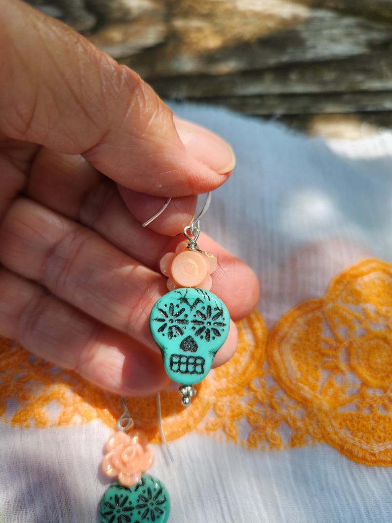 May include: Dangle earrings with turquoise skull beads and black floral patterns. Each earring has a peach flower bead and a silver hook. The earrings are displayed against a white and orange patterned fabric background.
