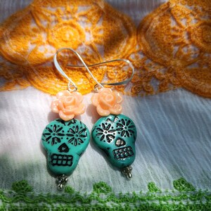 May include: A pair of turquoise skull earrings with silver accents and peach-coloured rose toppers. The skull earrings have black floral designs and are suspended from silver hooks. The earrings are displayed on a white cloth with orange and green embroidery.