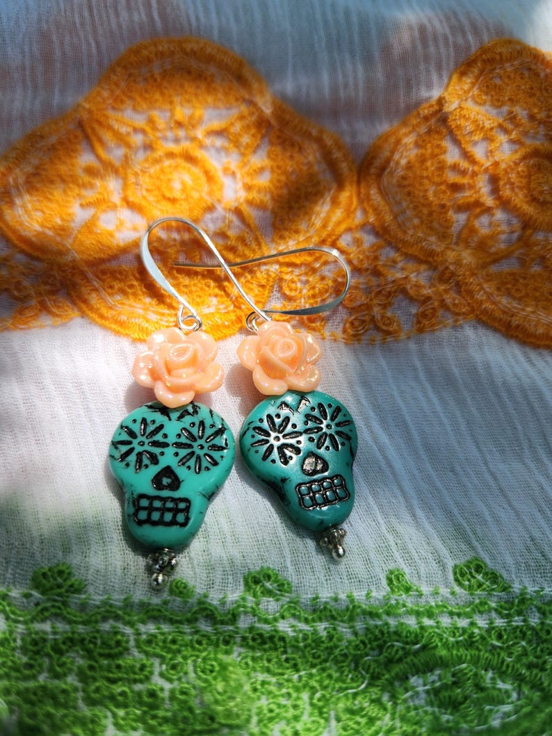 May include: A pair of turquoise skull earrings with silver accents and peach-coloured rose toppers. The skull earrings have black floral designs and are suspended from silver hooks. The earrings are displayed on a white cloth with orange and green embroidery.