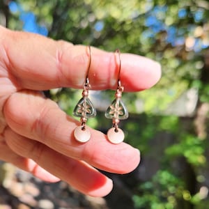 May include: A pair of copper-colored earrings. Each earring features a triangular, clear green glass bead with a floral design, a copper bead, and a small, round copper disc. The earrings are suspended from copper hooks.