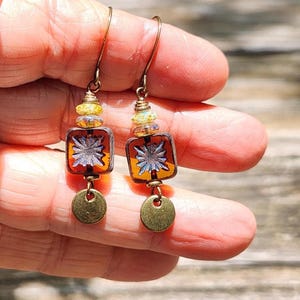 May include: A pair of drop earrings featuring a square amber-coloured glass bead with a white starburst pattern, accented by a small yellow faceted bead above and a round, brushed brass-toned metal disc dangling at the bottom on a wire hook.
