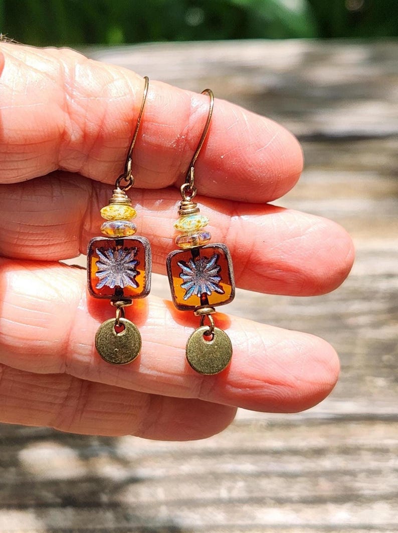 May include: A pair of drop earrings featuring a square amber-coloured glass bead with a white starburst pattern, accented by a small yellow faceted bead above and a round, brushed brass-toned metal disc dangling at the bottom on a wire hook.