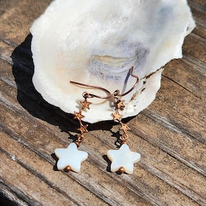 May include: Copper-colored star earrings with white star-shaped pendants. The earrings feature a series of small stars leading to a larger star pendant. The earrings are displayed on a white seashell against a wooden background.