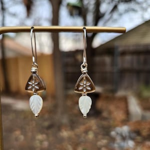 May include: A pair of dangle earrings. Each earring features a triangular, smoky quartz stone with a floral design, a white leaf-shaped mother-of-pearl, and a silver-colored metal hook. The earrings are displayed on a gold-colored metal bar.