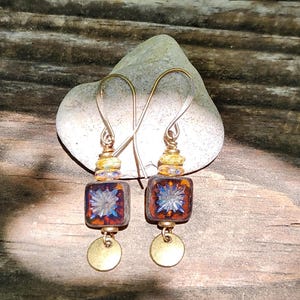 May include: A pair of dangle earrings featuring square, dark amber-coloured glass beads with a central blue starburst pattern. Each earring includes a small, round, metallic gold-toned disc at the base and a yellow, speckled glass bead above the square.