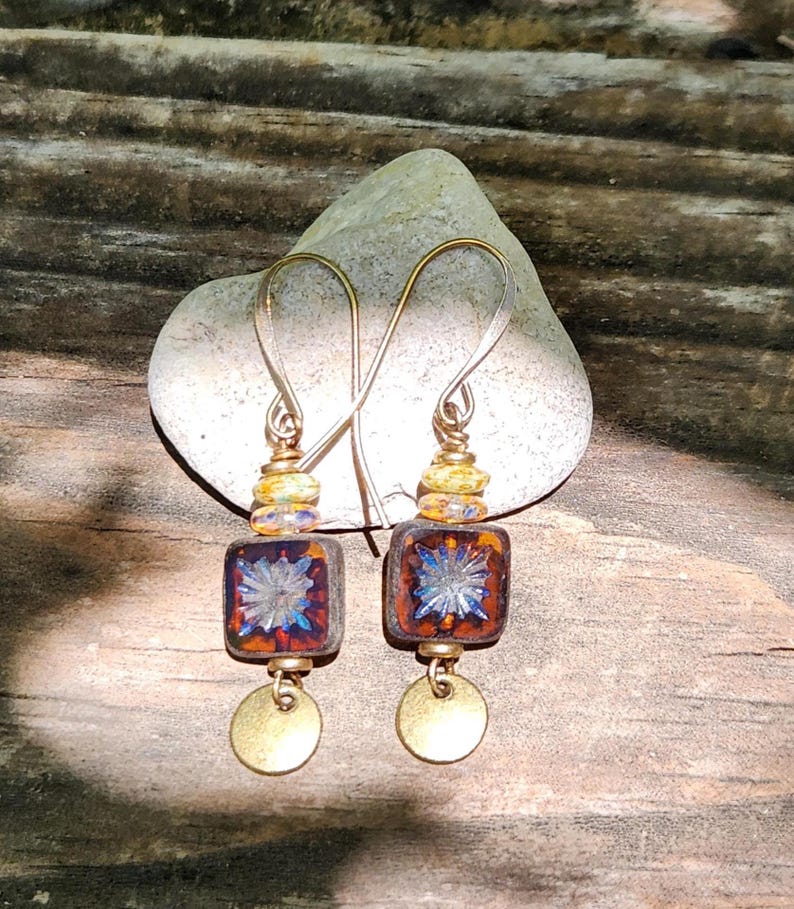 May include: A pair of dangle earrings featuring square, dark amber-coloured glass beads with a central blue starburst pattern. Each earring includes a small, round, metallic gold-toned disc at the base and a yellow, speckled glass bead above the square.