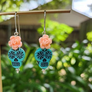 May include: Dangle earrings with turquoise skull-shaped beads and black detailing. Each earring has a peach-coloured flower accent and a silver hook. The earrings are displayed against a blurred green background.