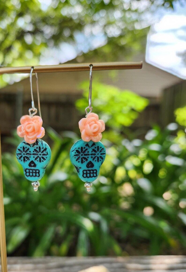 May include: Dangle earrings with turquoise skull-shaped beads and black detailing. Each earring has a peach-coloured flower accent and a silver hook. The earrings are displayed against a blurred green background.