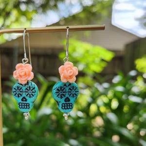 May include: Dangle earrings with turquoise skull beads, black floral patterns, and peach flower accents. The earrings hang from silver hooks. The design is inspired by the Day of the Dead, a Mexican holiday.