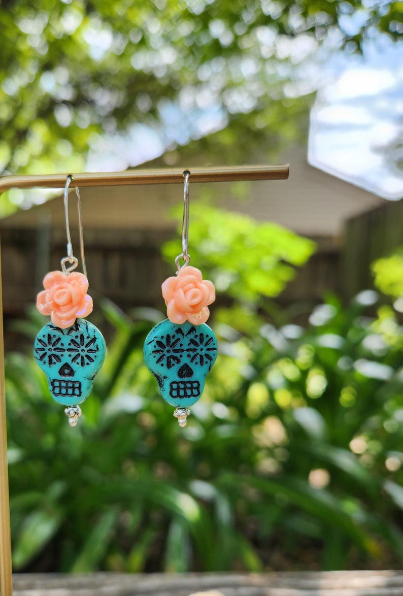 May include: Dangle earrings with turquoise skull beads, black floral patterns, and peach flower accents. The earrings hang from silver hooks. The design is inspired by the Day of the Dead, a Mexican holiday.