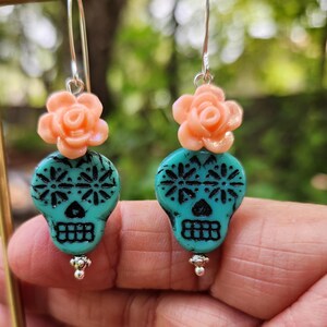May include: Dangle earrings with turquoise skull beads and black detailing, topped with peach rose accents. The earrings have silver hooks and small silver beads. The background is a blurred green. The earrings are a unique accessory.