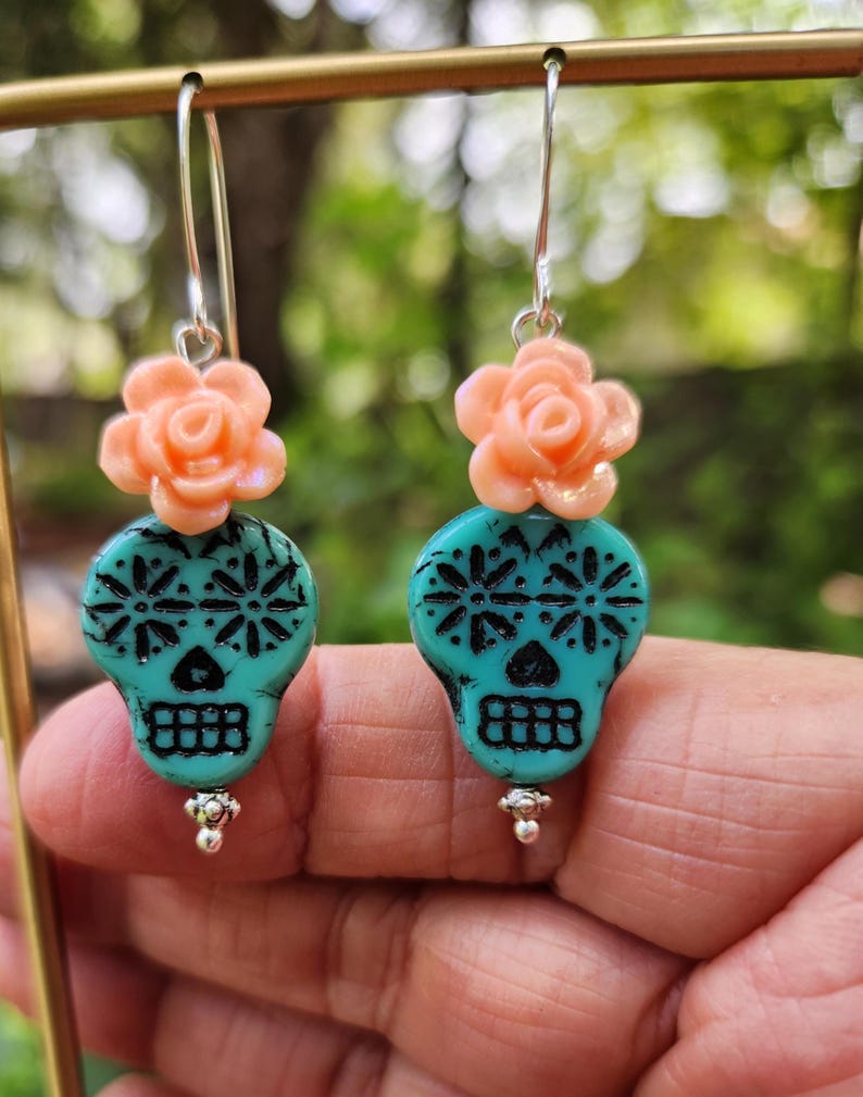 May include: Dangle earrings with turquoise skull beads and black detailing, topped with peach rose accents. The earrings have silver hooks and small silver beads. The background is a blurred green. The earrings are a unique accessory.