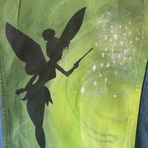 May include: A green denim jacket featuring a black silhouette of Tinkerbell holding a wand and sprinkling glitter. The glitter is white and resembles stars.