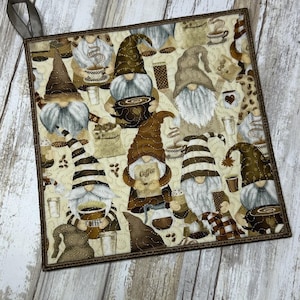 May include: Square quilted potholder featuring a repeating pattern of coffee-loving gnomes in brown and beige tones. The gnomes are holding coffee cups and wearing hats. The potholder has a brown border and a fabric loop for hanging.