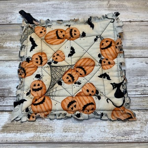 May include: A white quilted pot holder with a Halloween theme. It features orange and black pumpkins, bats, and a spider web. The pot holder has a frayed edge.