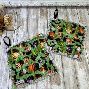 May include: Two green fabric pot holders with a white frayed edge. The pot holders feature a pattern of cupcakes with various toppings, bats, and spiderwebs. The text "Bakery Craft" is visible on one of the pot holders.