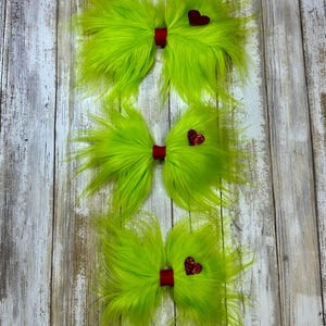 Peut inclure: Trois nœuds papillon en fourrure verte avec des cœurs rouges pailletés. Les nœuds papillon sont disposés verticalement, le plus grand en haut et le plus petit en bas.