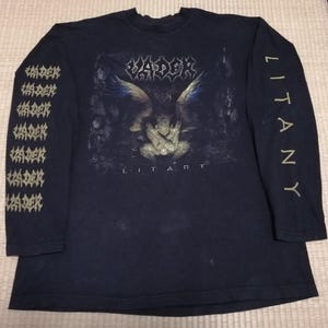 May include: Black long-sleeve shirt featuring the band name "VADER" above a graphic of an angel. The sleeves display "VADER" and "LITANY" in gold lettering. A dark, vintage-style band tee.
