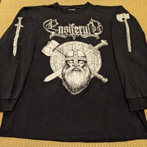 May include: Black long-sleeve shirt featuring the band name "Ensiferum" in white gothic lettering. The shirt has a graphic of a bearded Viking with a helmet, shield, and crossed swords. A sword and axe are printed on the sleeves.