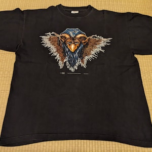 May include: Black t-shirt with a graphic print of a blue and brown creature with wings. The creature has a beard and a crown.
