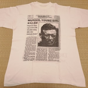 May include: White t-shirt with a black and white newspaper print featuring a headline that reads "Murder, Young Girl Killed." The newspaper article includes a black and white photo of a man.