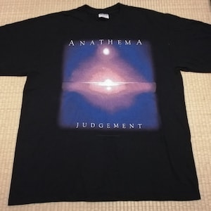 May include: Black t-shirt featuring the band name "ANATHEMA" above a graphic with a purple and blue gradient design. The word "JUDGEMENT" is printed below the graphic. The shirt is made of a soft material.