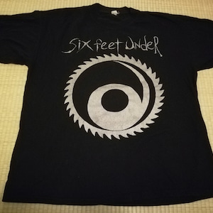 May include: Black t-shirt with a white graphic of a circular saw blade with a stylized "6" inside. The text "Six Feet Under" is printed above the graphic.