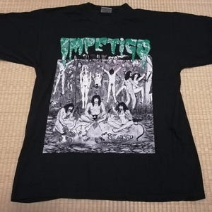 May include: Black t-shirt with a graphic print featuring the word "Impettigo" in green, above a black and white illustration of figures in a forest setting. The shirt has a crew neck and short sleeves.