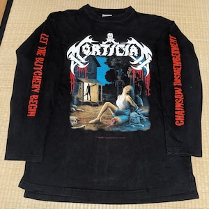 Puede incluir: Camiseta negra de manga larga con un gráfico que presenta el nombre de la banda "Mortician" en blanco, una calavera y una escena de terror. Las mangas tienen las frases "Let the Butchery Begin" y "Chainsaw Dismemberment" en rojo.