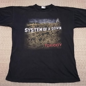 May include: Black t-shirt featuring the band name "SYSTEM OF A DOWN" in white block letters above a landscape graphic. Below the graphic, the word "TOXICITY" is printed in red. The shirt is made of a soft material.