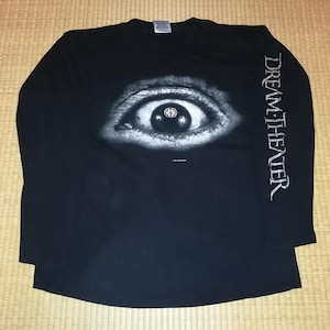 May include: Black long-sleeve shirt featuring a large, detailed eye graphic on the front. The eye has a grayscale design with a band logo in the center. The band name, DREAM THEATER, is printed vertically on the right sleeve.