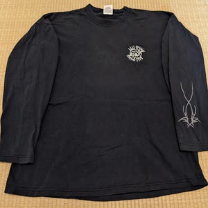 Op de afbeelding: Zwart langarm T-shirt met een witte afbeelding van een schedel met vleugels en de tekst "All Rise" op de voorkant. De rechtermouw heeft een witte afbeelding van een gestileerd vlammenontwerp.